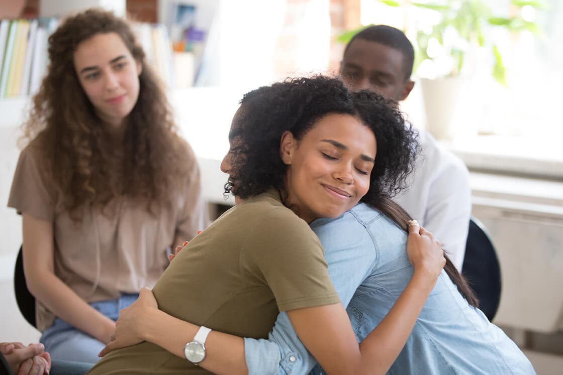 Man and woman hugging in a therapy session. Others are looking and smiling.