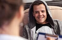 Youth boy hearing a grey hoodie and sitting in a chair. Seen from behind a woman, blurry in the foreground